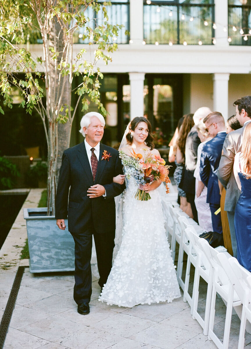 A bride is escorted to the aisle of her art-inspired wedding ceremony on the arm of her father, carrying a colorful bouquet in her other hand.