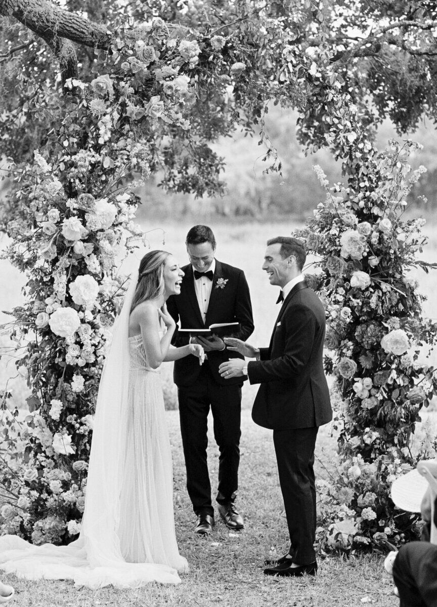A bride and groom laugh during their elegant spring wedding ceremony, which took place in front of a floral arch.